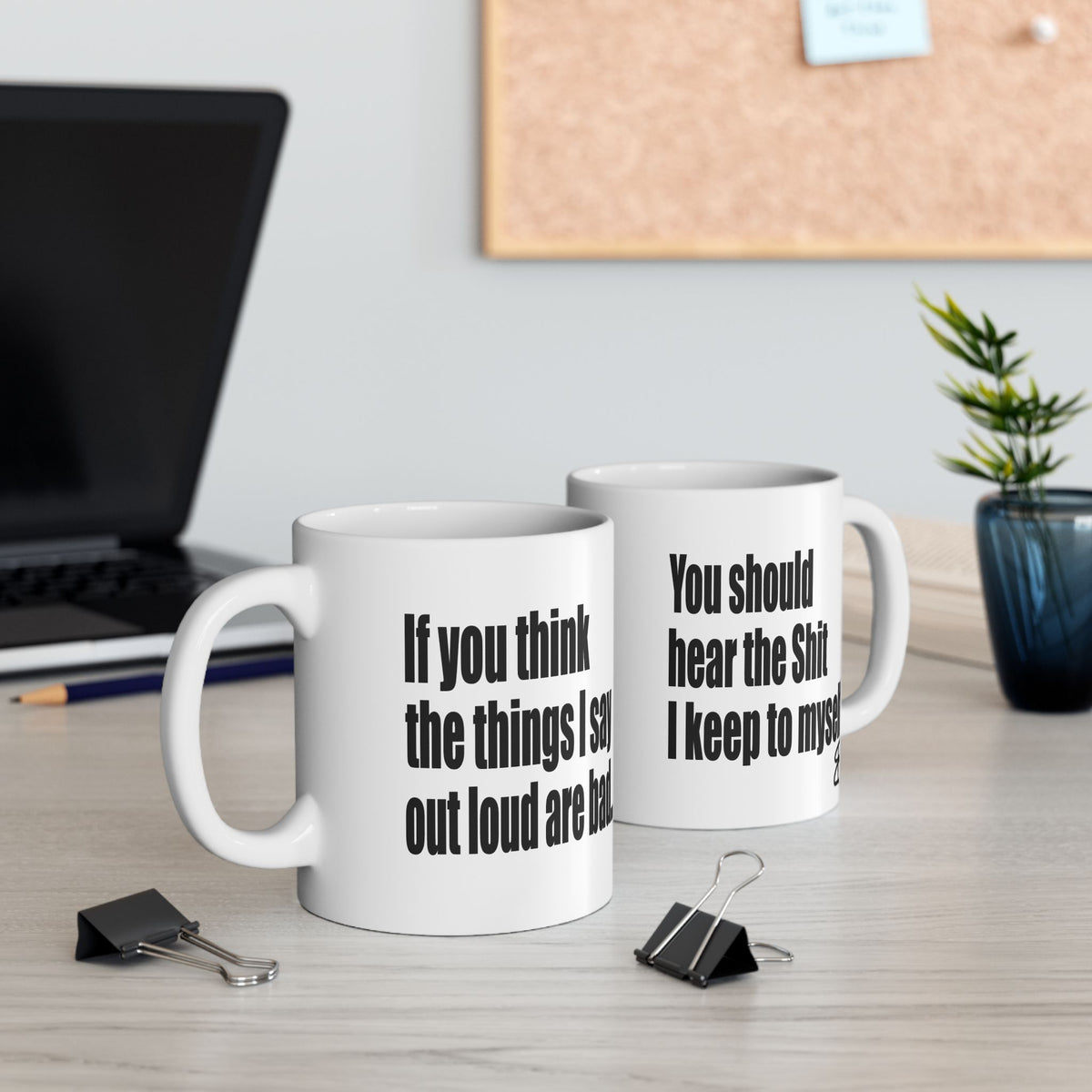 Two white ceramic mugs with black printed text on a light wood desk, styled with office props like binder clips, a laptop, and a small plant in the background.