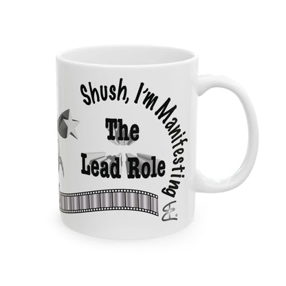 A white ceramic mug with a black printed graphic that reads 'Shush, I'm Manifesting The Lead Role' with a film strip design, shown on a plain white background.