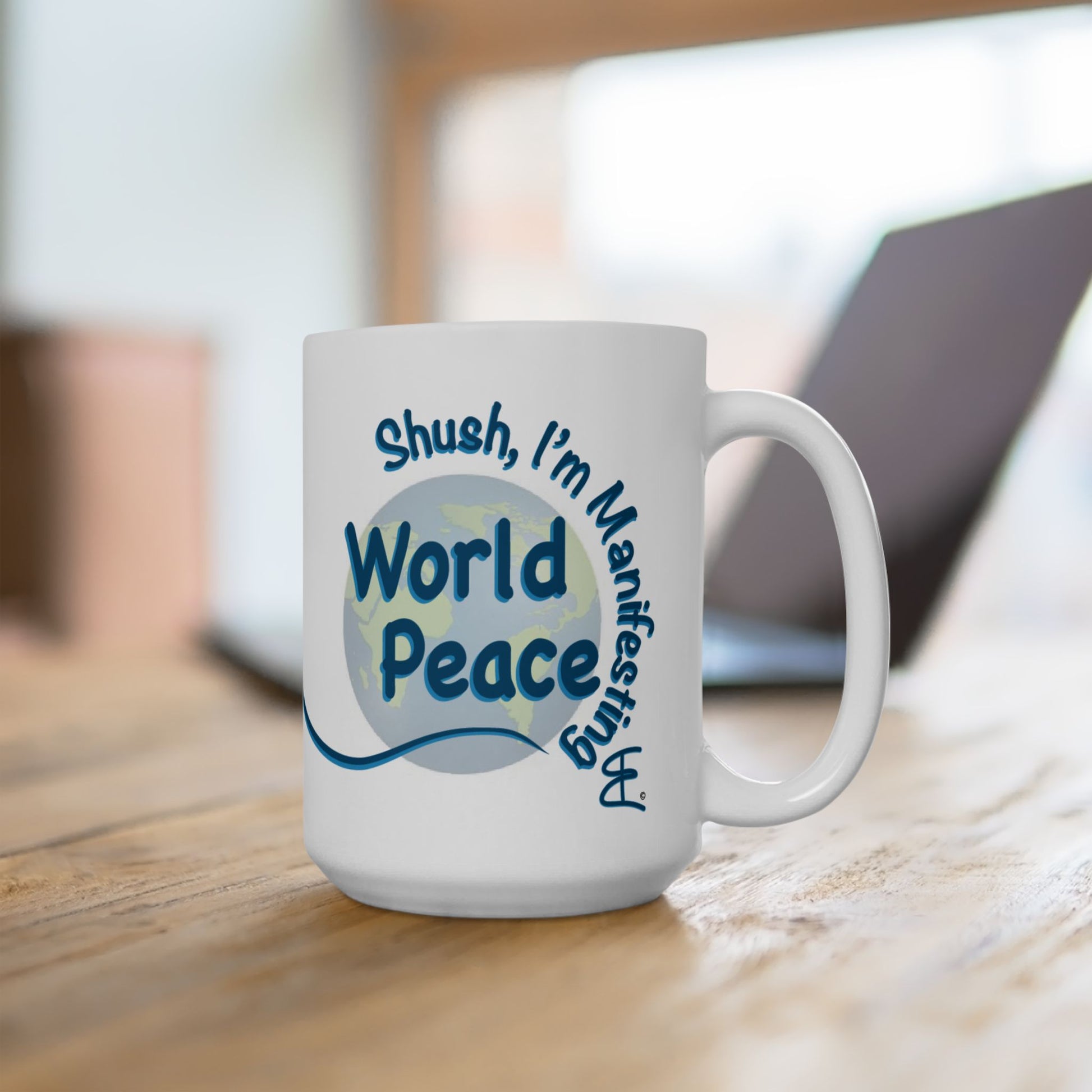 A white ceramic coffee mug with a blue and light blue graphic that reads 'Shush, I'm Manifesting World Peace', placed on a wooden table with a blurred laptop in the background.