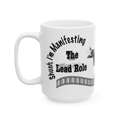 A white ceramic mug with a black printed graphic that reads 'Shush, I'm Manifesting The Lead Role' with a film strip design, shown on a plain white background.