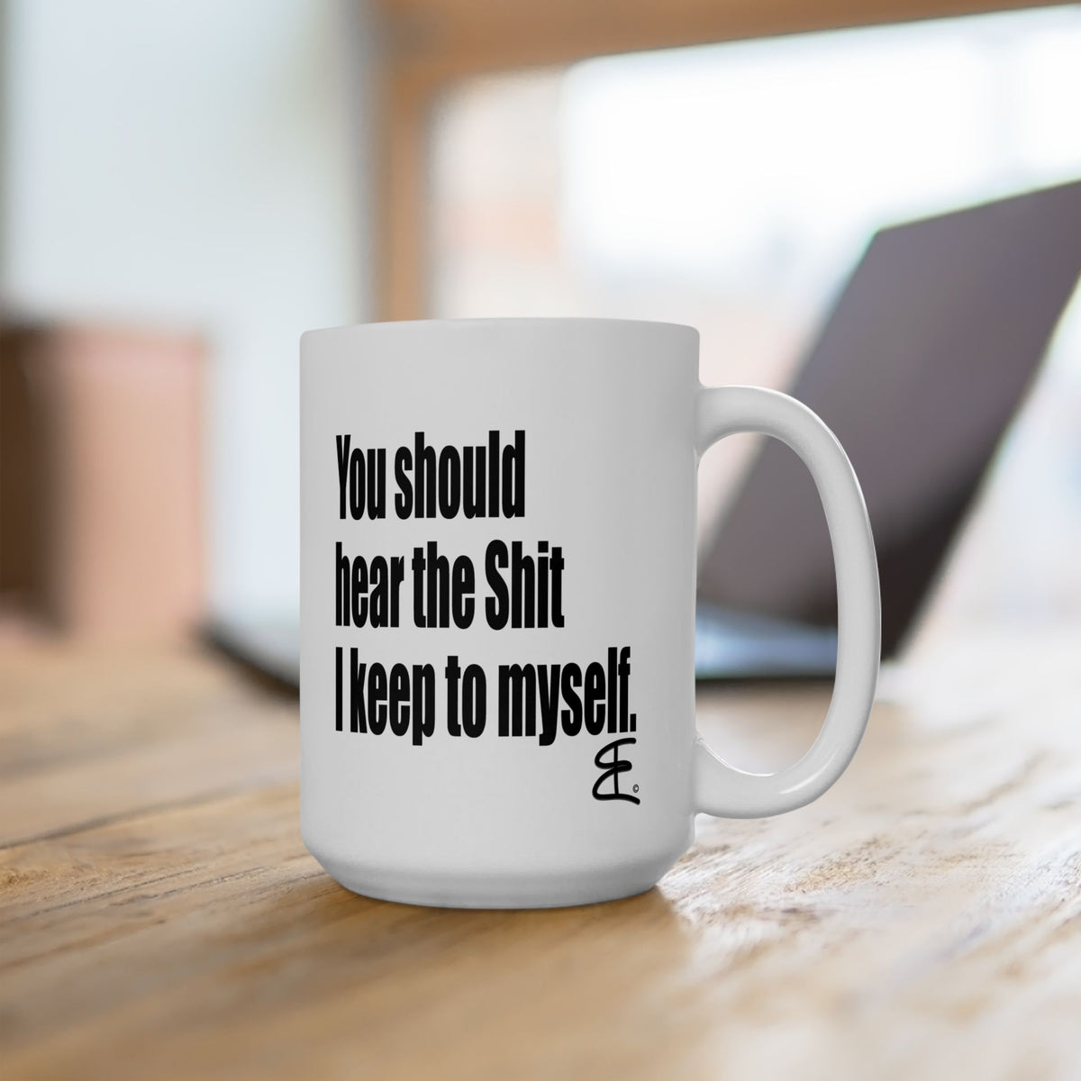 A white ceramic coffee mug with bold black text reading 'You should hear the Shit I keep to myself.' sitting on a wooden table with a blurred laptop in the background.
