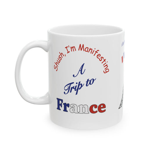 A white ceramic coffee mug with left handle, featuring a printed design that reads 'Shush, I'm Manifesting A Trip to France' in red, blue  and white lettering.