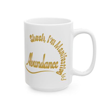 A white ceramic coffee mug with gold cursive text that reads 'Shush, I'm Manifesting Abundance' on a clean white background.