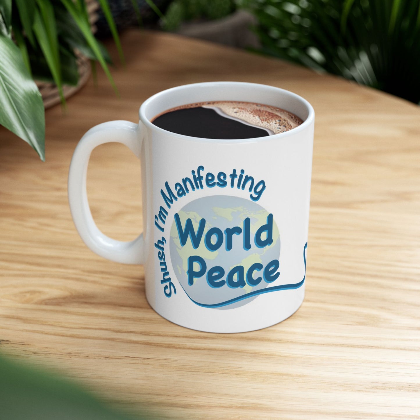 A white ceramic coffee mug with a blue and light blue graphic that reads 'Shush, I'm Manifesting World Peace' sitting on a light wood table with blurred green plants in the background.