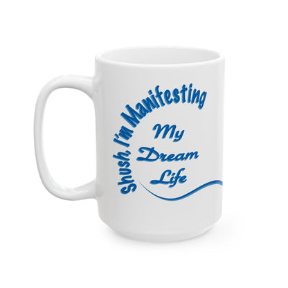 A white ceramic coffee mug with a blue printed graphic that reads 'shush I'm Manifesting My Dream Life' on a clean white background.