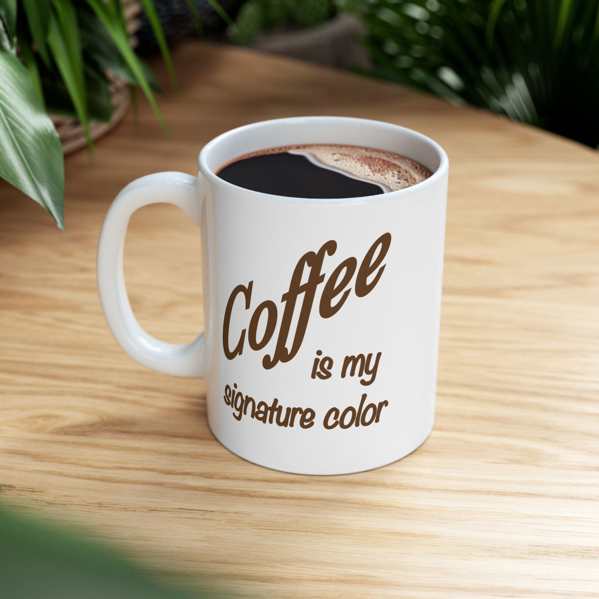 A white mug with 'Coffee is my signature color' printed in brown, filled with black coffee sitting a light wooden table.