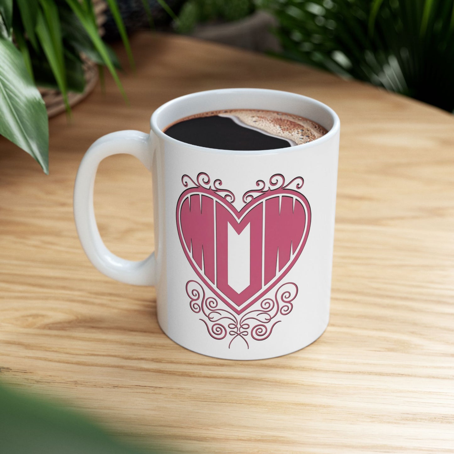 White ceramic mug featuring "Mom" printed in pink text inside a heart with decorative filigree, on a wooden desk.