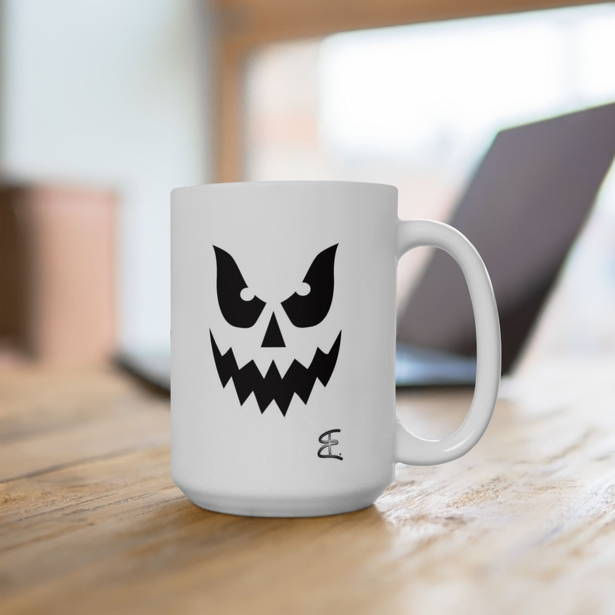 A white ceramic mug with a black ghost face graphic, sitting on a wooden table with a blurred laptop in the background.