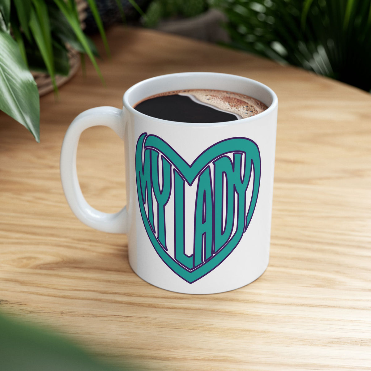 A white ceramic coffee mug filled with black coffee sits on a light wood table. The mug features a graphic of a teal heart outline with the words 'MY LADY' in stylized heart shape.
