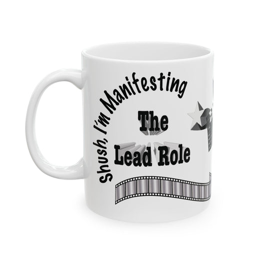 A white ceramic mug with a black printed graphic that reads 'Shush, I'm Manifesting The Lead Role' with a film strip design, shown on a plain white background.