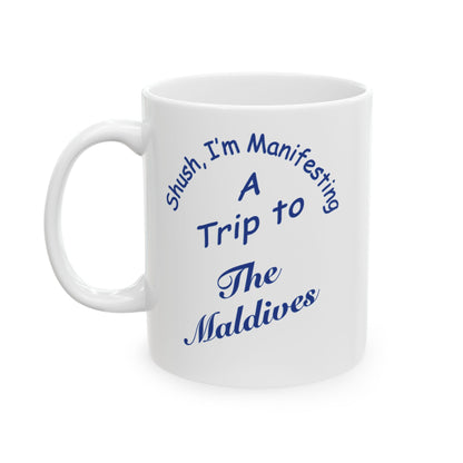 A white ceramic coffee mug with blue printed text that reads 'Shush, I'm Manifesting A Trip to The Maldives' on a clean white background.