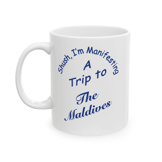 A white ceramic coffee mug with blue printed text that reads 'Shush, I'm Manifesting A Trip to The Maldives' on a clean white background.