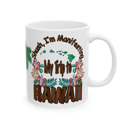 A white ceramic coffee mug featuring a colorful graphic design with the text 'Shush, I'm Manifesting My Trip to HAWAII' and tropical floral illustrations.