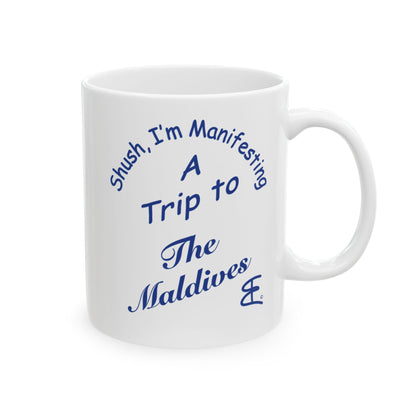 A white ceramic mug with blue printed text that reads 'Shush, I'm Manifesting A Trip to The Maldives' on a clean white background.