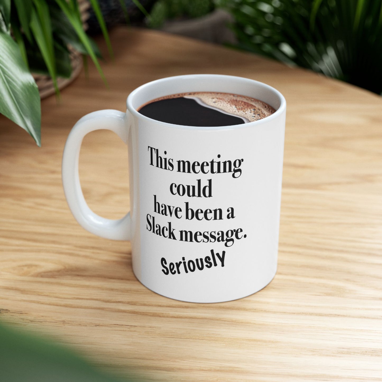 A white ceramic mug with text "This Meeting Could Have Been A Slack Message"  on a wooden desk with plants in background.