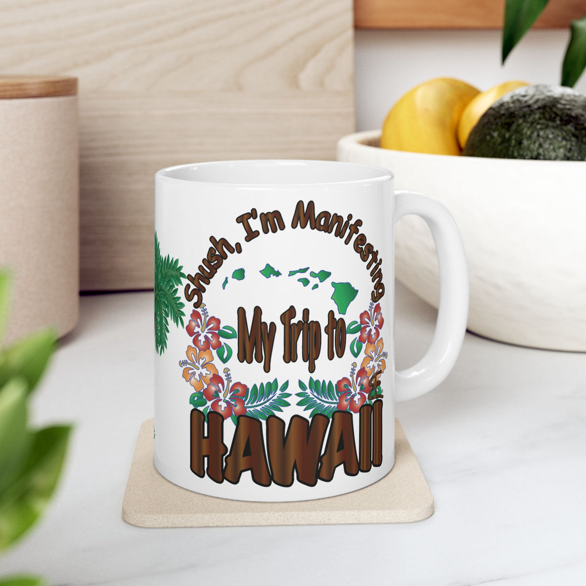 A white ceramic mug with a colorful tropical Hawaiian design and the text 'Shush, I'm Manifesting My Trip to HAWAII' sitting on a light wood coaster on a white marble countertop.