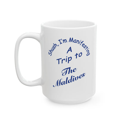 A white ceramic coffee mug with a blue printed text design that reads 'Shush, I'm Manifesting A Trip to The Maldives' on a clean white background.
