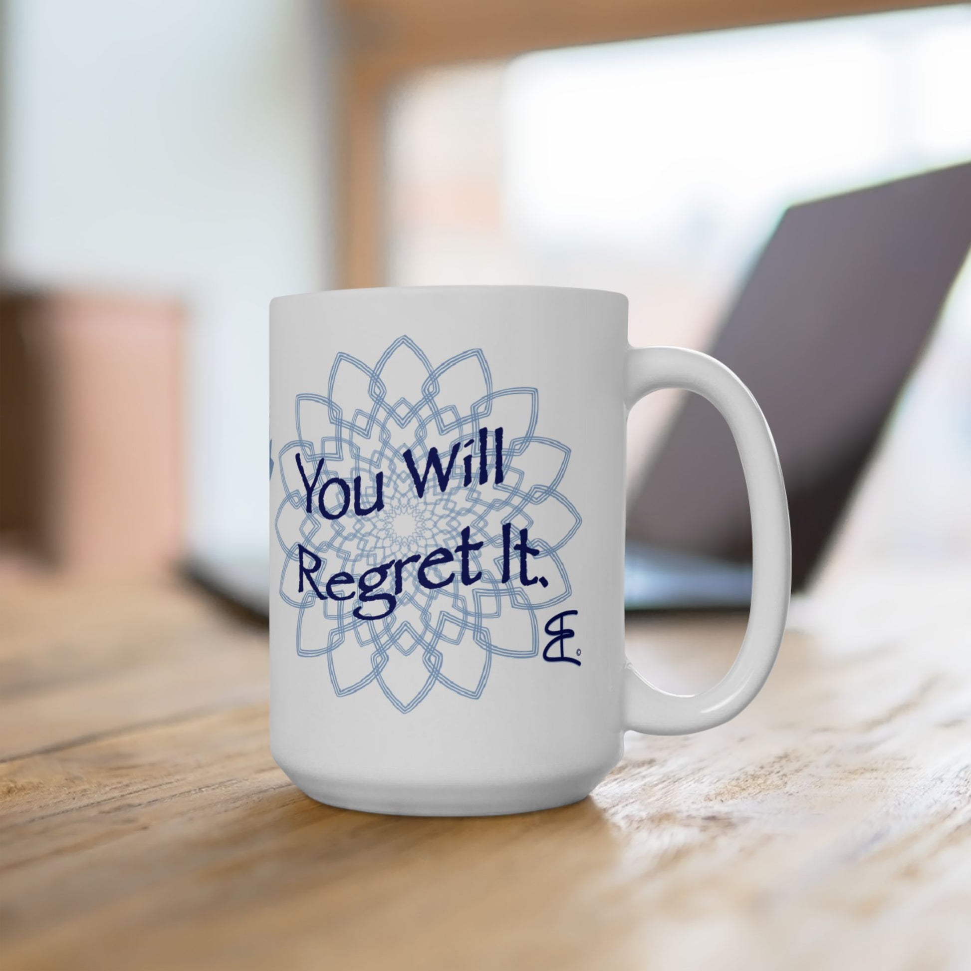 White ceramic mug with blue mandala artwork and "You Will Regret It." on a wooden table beside a blurred laptop.