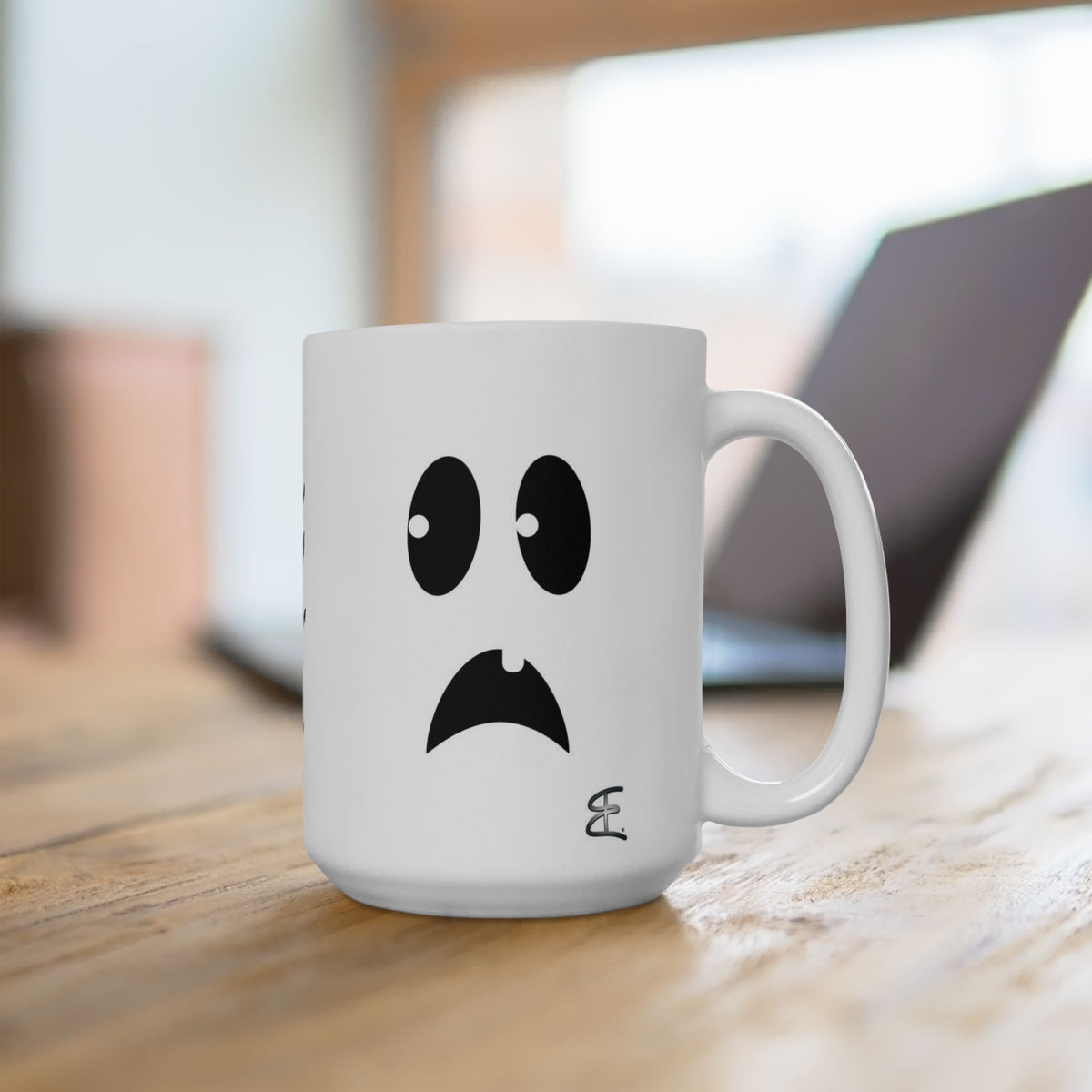 A white ceramic coffee mug with a black cartoon ghost face printed on it, sitting on a wooden table with a blurred laptop in the background.