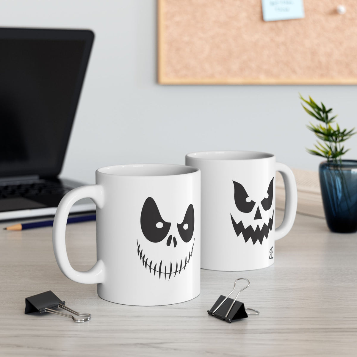 Two white mugs with spooky ghost faces on a desk with office items.