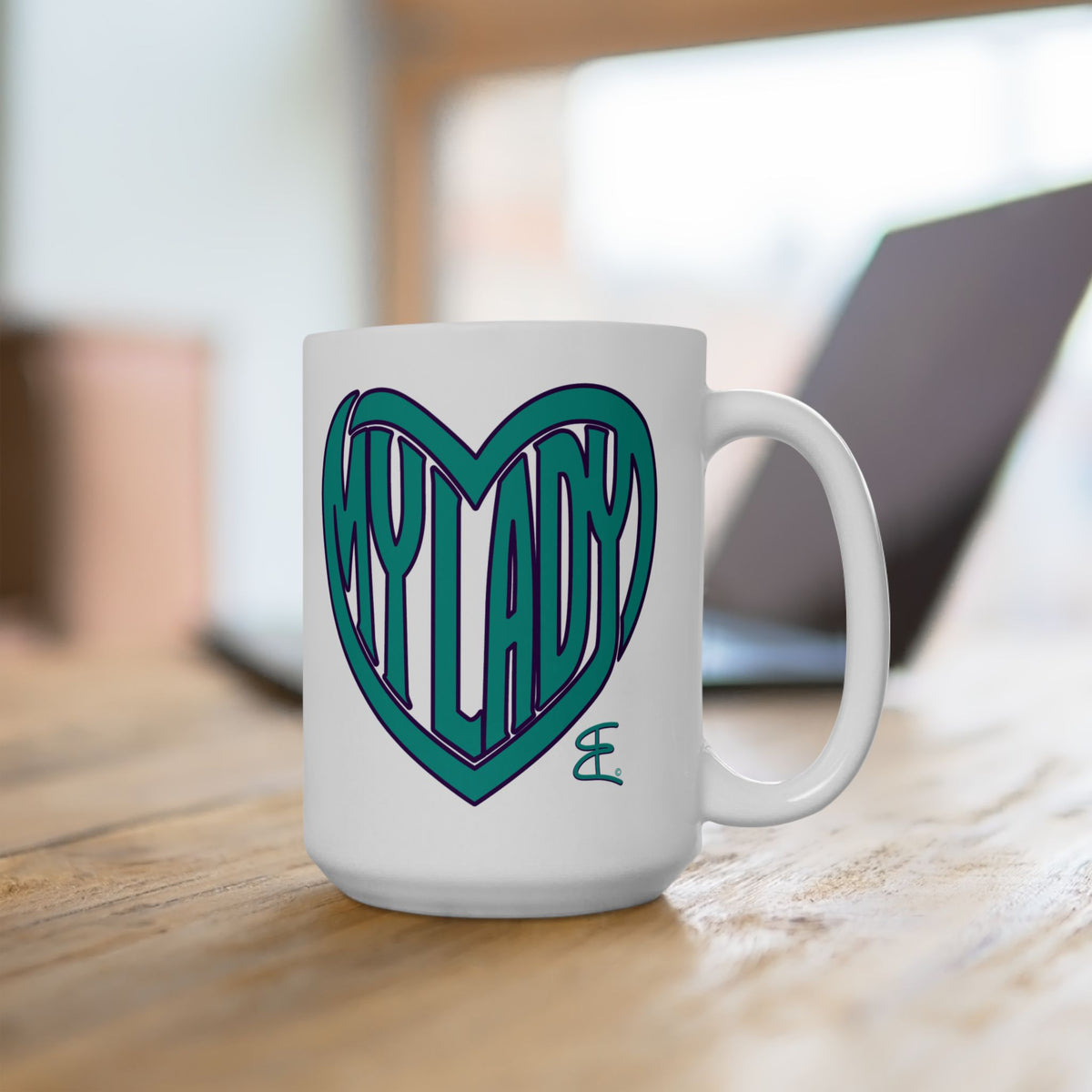 A white ceramic coffee mug with a teal heart graphic containing the text 'MY LADY', sitting on a wooden table with a blurred laptop in the background.