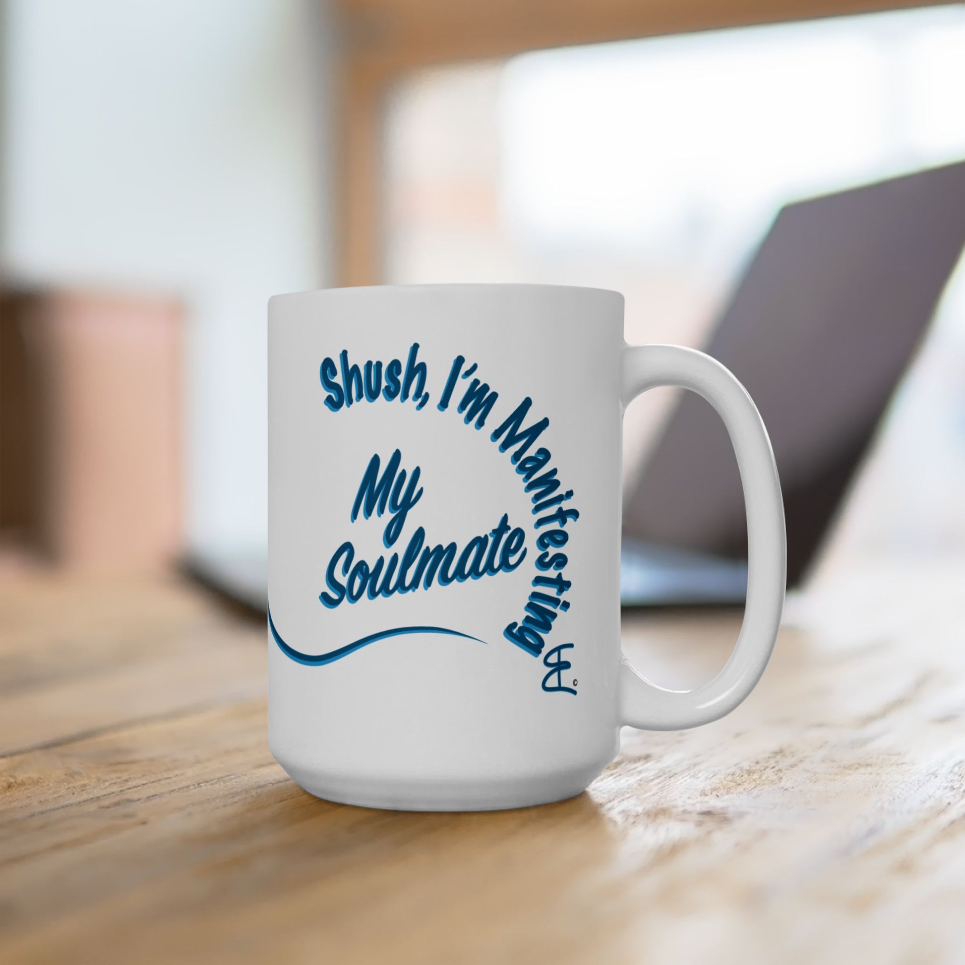A white ceramic coffee mug with blue text reading 'Shush, I'm Manifesting My Soulmate' sitting on a wooden table with a blurred laptop in the background.