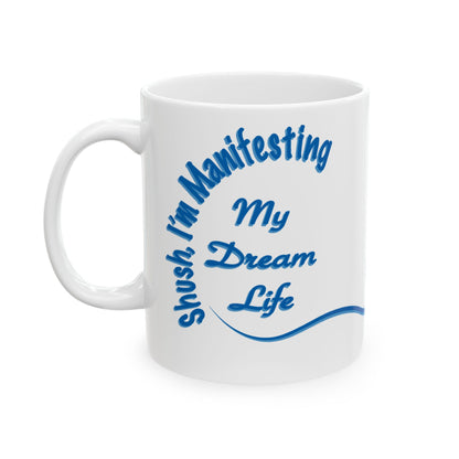 A white ceramic coffee mug with a blue printed graphic that reads 'shush, I'm Manifesting My Dream Life' in a stylized script font, set against a plain white background.