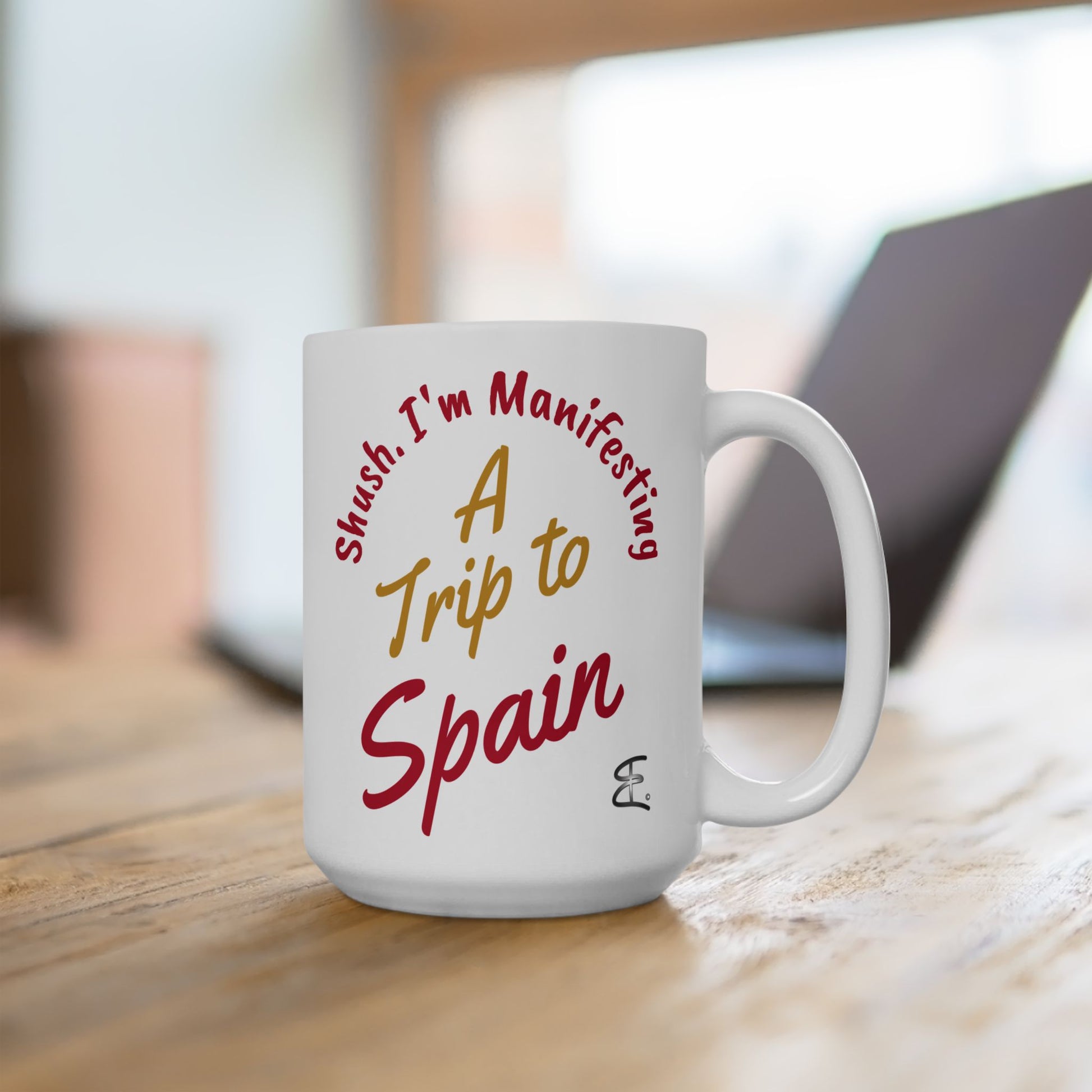 'Shush I'm Manifesting A Trip to Spain' 15oz ceramic mug lifestyle picture on wooden desk with laptop in background.