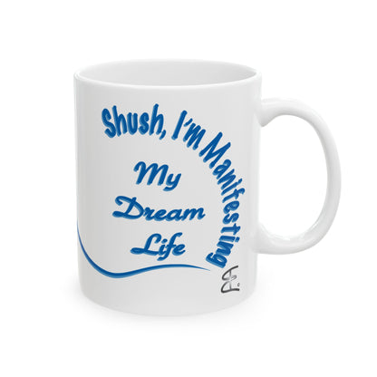 A white ceramic mug with blue text reading 'Shush, I'm Manifesting My Dream Life' and a blue curved line, shown on a plain white background.