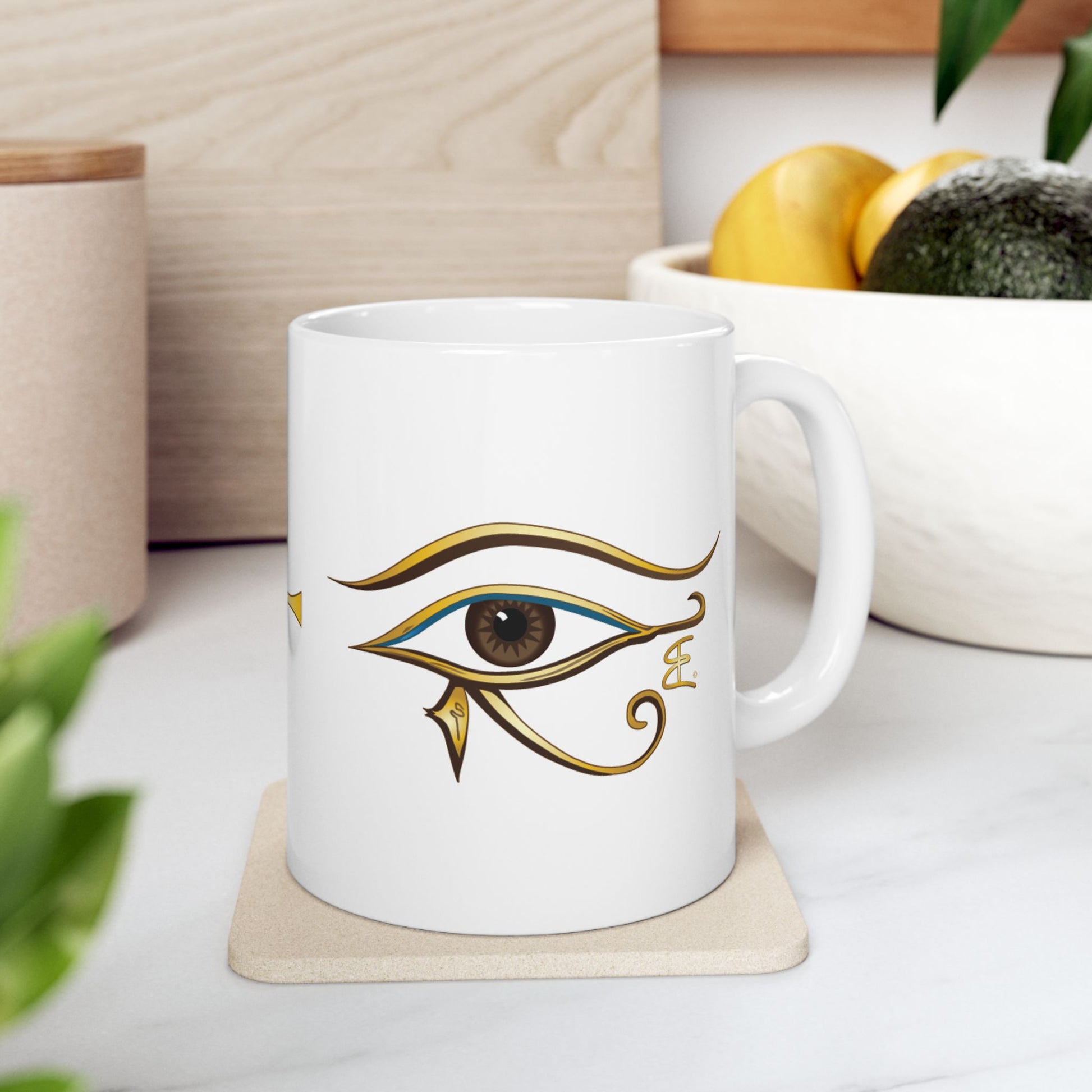 A white ceramic mug featuring a gold and green Egyptian Eye of Horus design, resting on a light wood coaster on a white marble countertop with a blurred kitchen background.