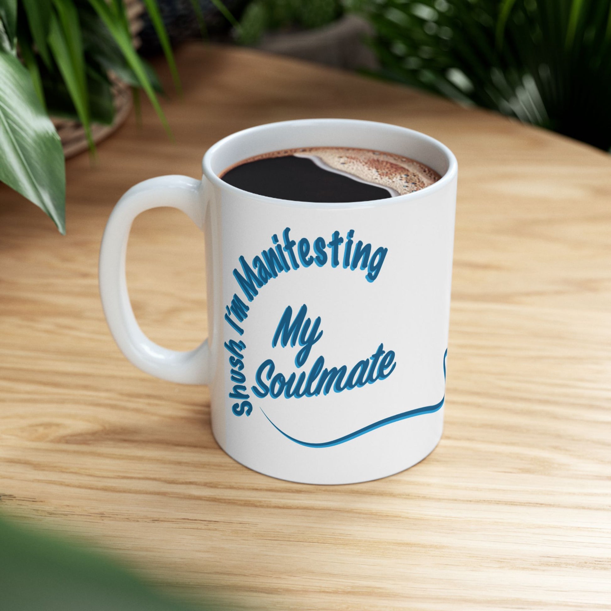 A white ceramic coffee mug with blue text reading 'Shush I'm Manifesting My Soulmate' while filled with black coffee, sitting on a light wood table with blurred green plants in the background.