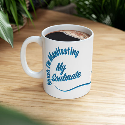 A white ceramic coffee mug with blue text reading 'Shush I'm Manifesting My Soulmate' while filled with black coffee, sitting on a light wood table with blurred green plants in the background.