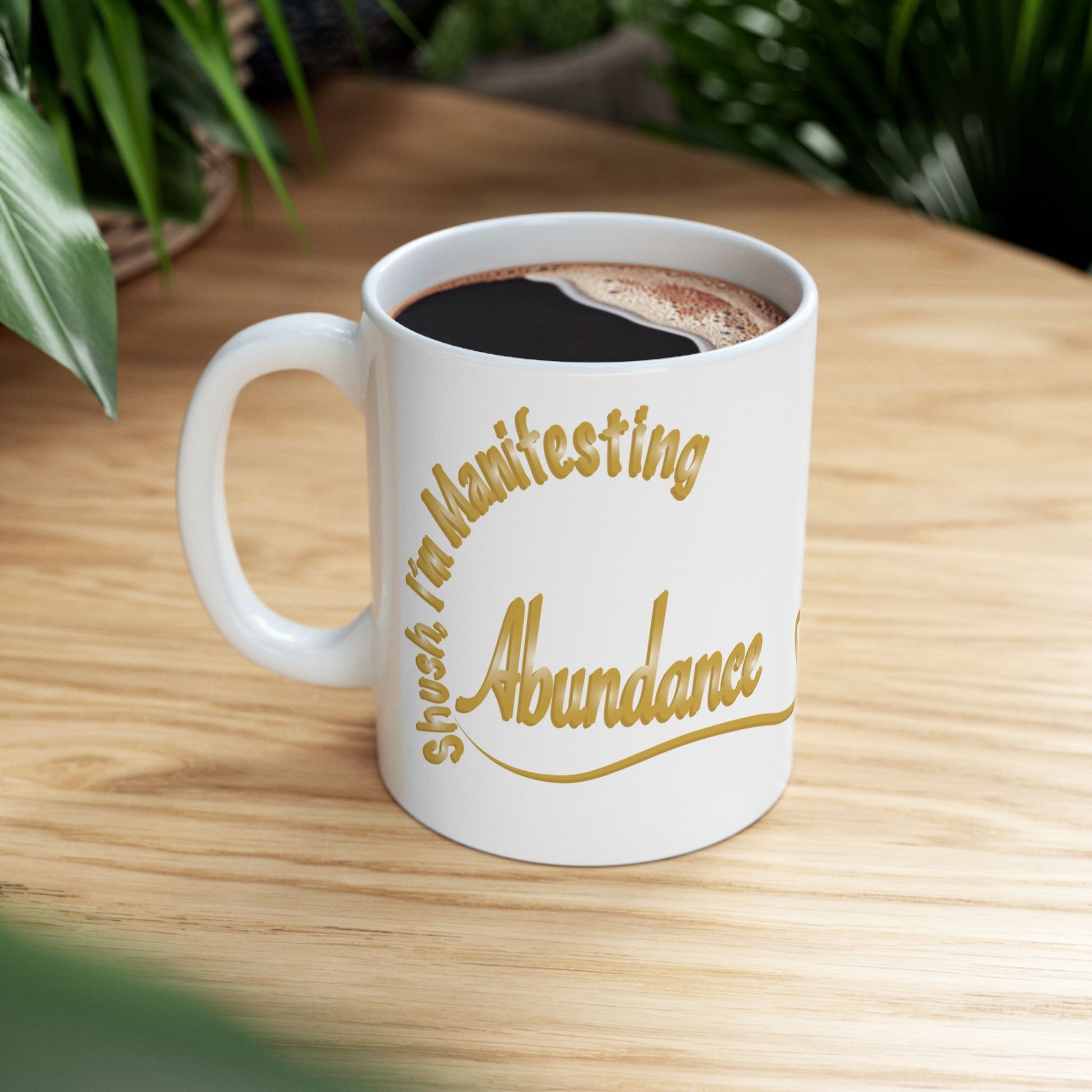 A white ceramic coffee mug with gold text reading 'Shush, I'm Manifesting Abundance', filled with black coffee and set on a light wooden table with blurred green plants in the background.