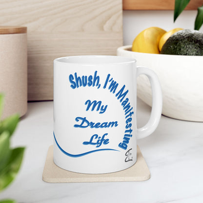 A white ceramic coffee mug with blue text reading 'Shush, I'm Manifesting My Dream Life' sitting on a light wood coaster on a kitchen counter.