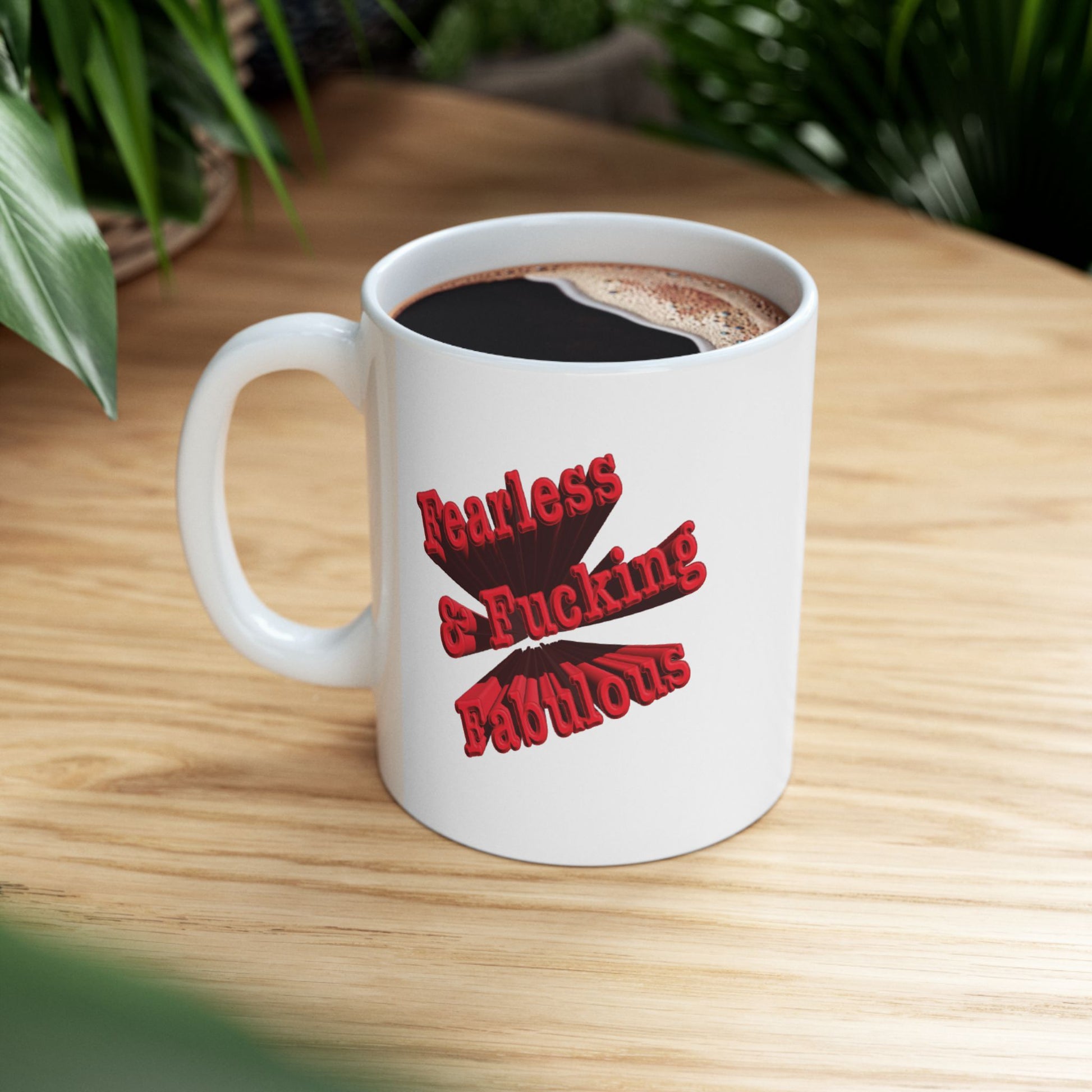 A white ceramic mug with a red graphic text design that reads 'Fearless & Fucking Fabulous' sitting on light wooden desk.