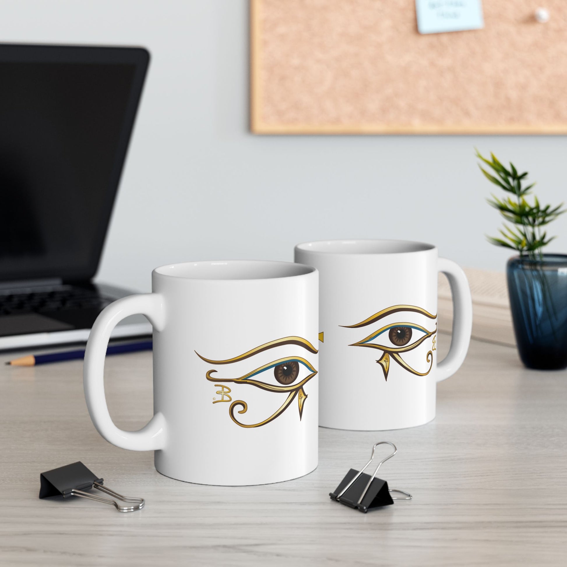 Two white mugs showing the eye of Horus is on both sides of the mug, on a desk with office items.