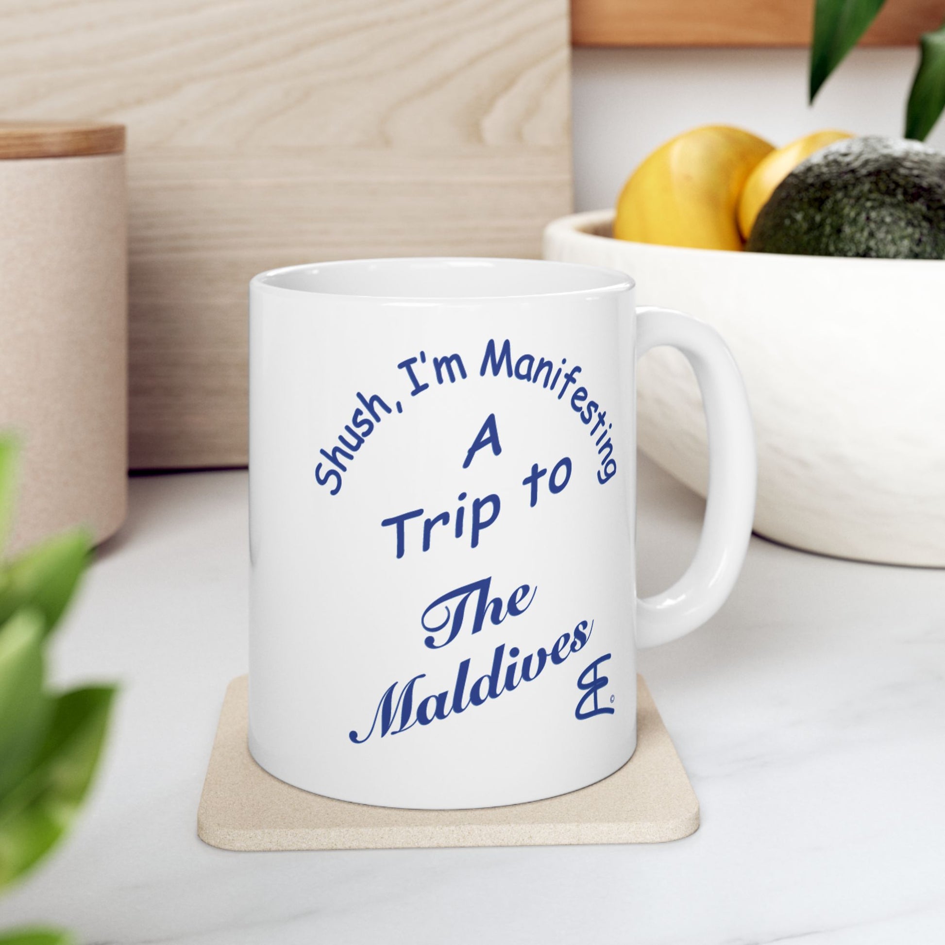 A white ceramic mug with blue text reading 'Shush, I'm Manifesting A Trip to The Maldives' sits on a light wood coaster on a kitchen counter, with a bowl of fruit and plants blurred in the background.