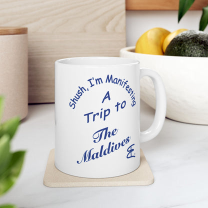 A white ceramic mug with blue text reading 'Shush, I'm Manifesting A Trip to The Maldives' sits on a light wood coaster on a kitchen counter, with a bowl of fruit and plants blurred in the background.