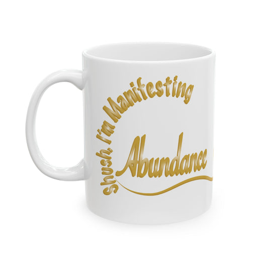 A white ceramic mug with a gold printed text design that reads 'Shush I'm Manifesting Abundance' on a clean white background.