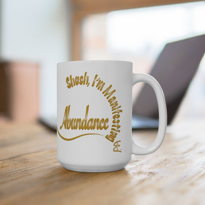 White mug with gold text that says, 'Shush I'm manifesting Abundance' on a wooden surface.